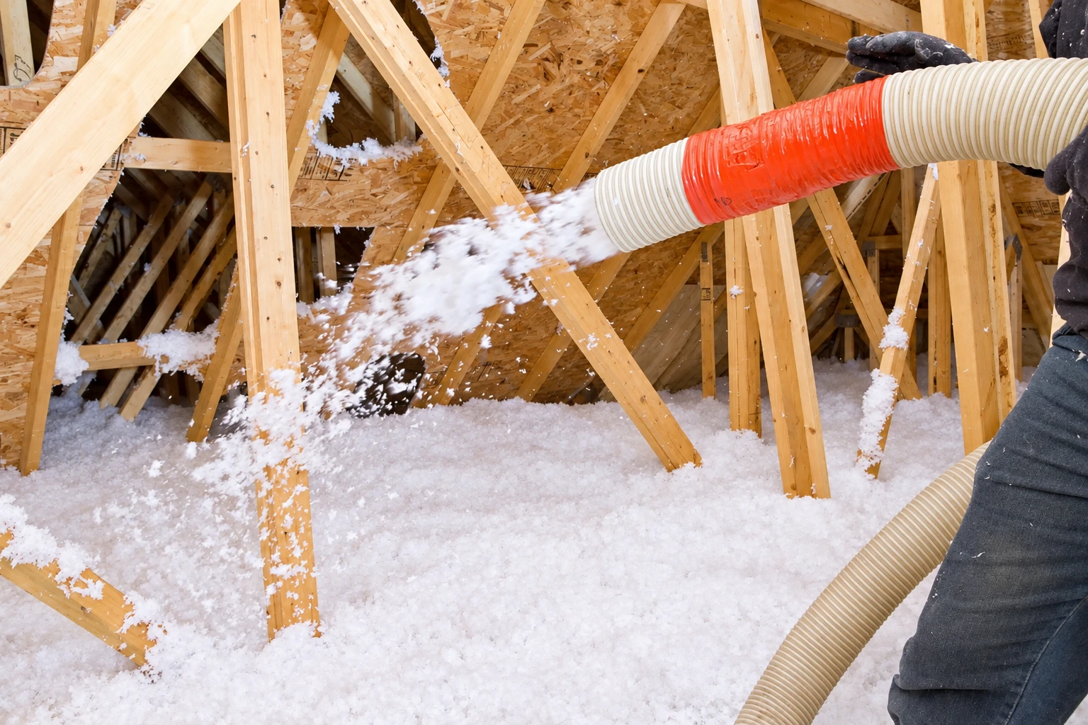A hose blows white loose-fill insulation across an attic floor among exposed wooden roof trusses.