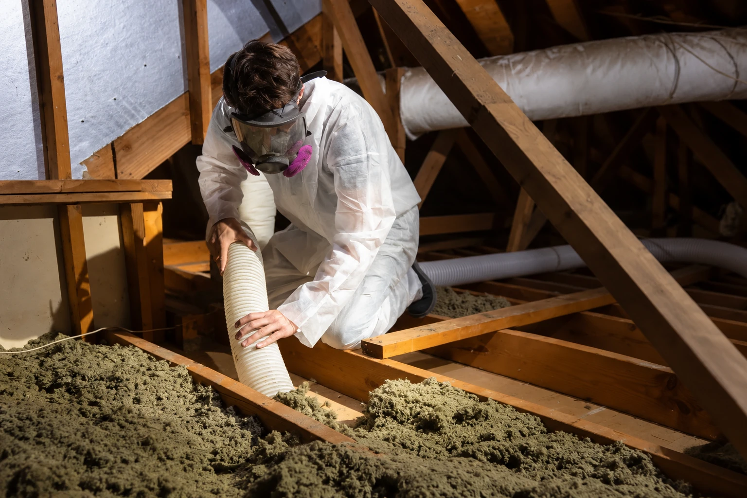 A worker in protective coveralls and a respirator uses a hose to remove loose insulation from attic floor joists.