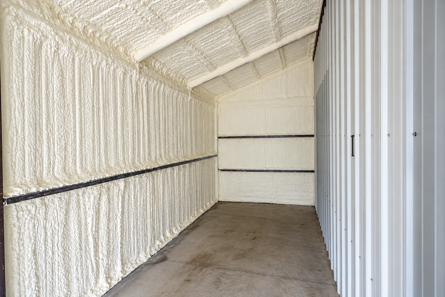 A narrow storage unit has cream spray foam covering its walls and sloped ceiling beside a concrete floor.