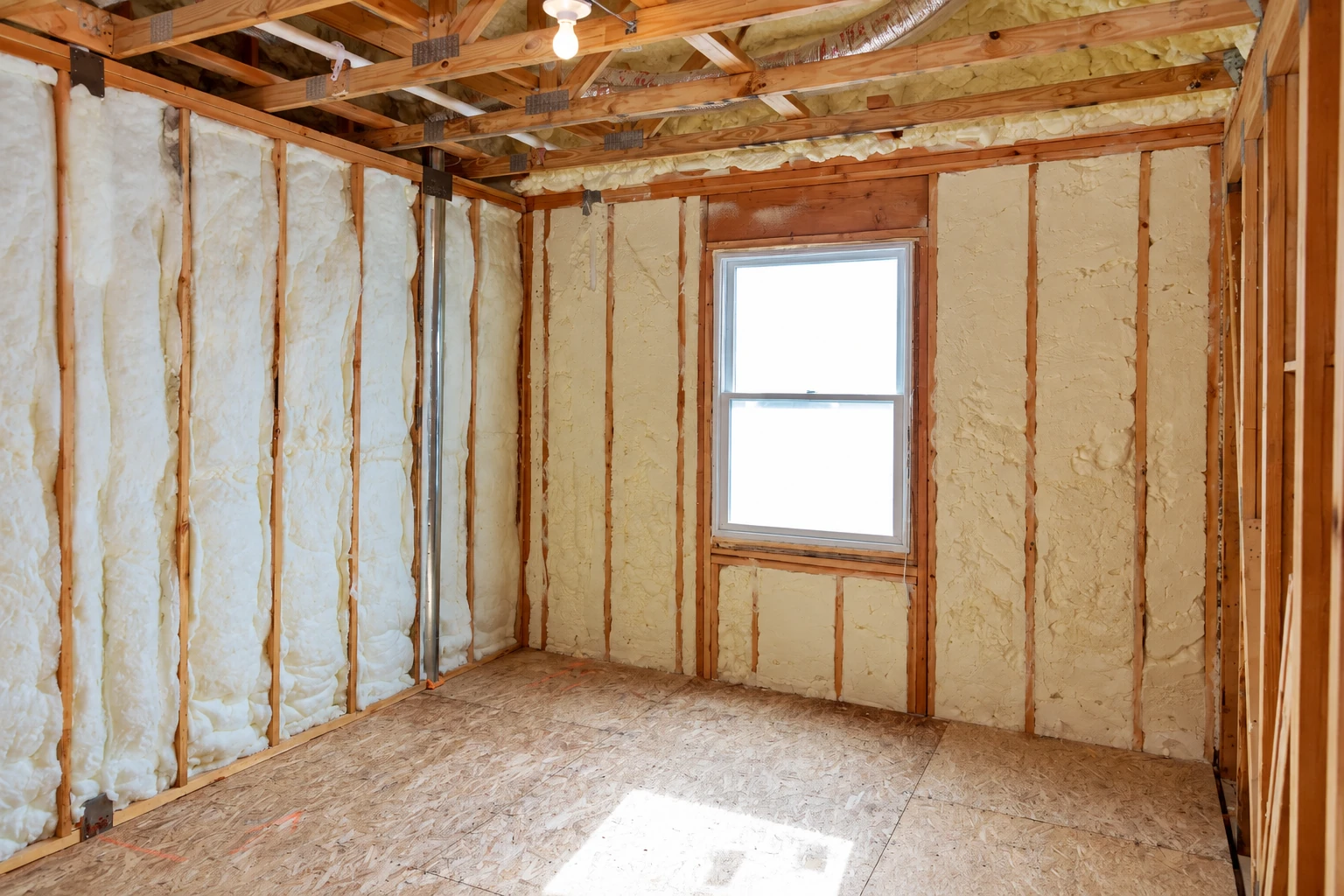 Cream-colored spray foam fills the wall cavities of a framed room with an exposed subfloor and a central window.