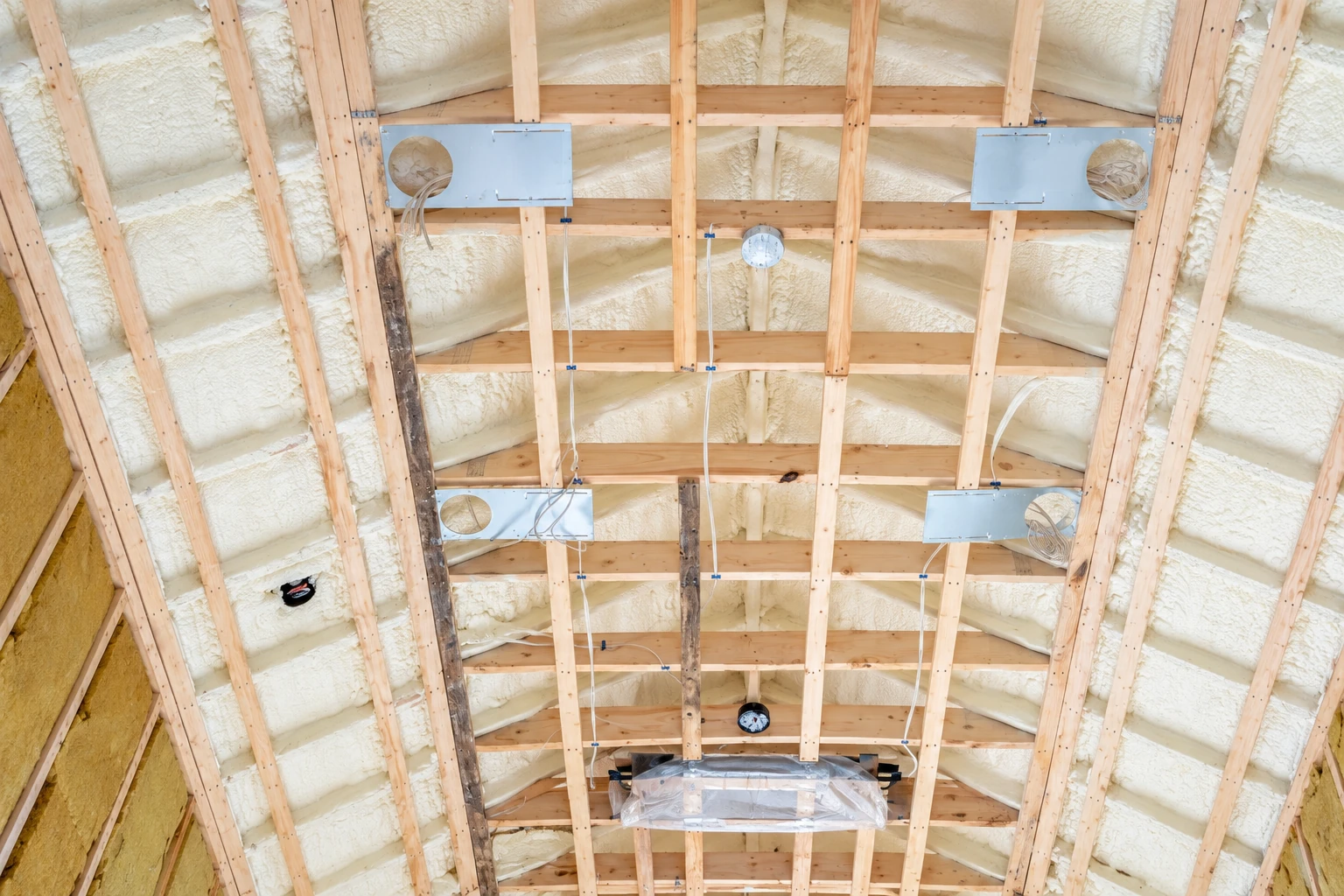 Exposed roof framing is coated with cream spray foam, with ducts and wiring visible between the joists.